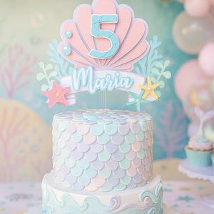 May include: A two-tiered cake with a mermaid theme. The cake is decorated with pastel colors and a scale pattern. A topper features the number 5 and the name "Maria". The bottom tier has a wave design.
