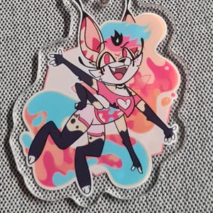 May include: A clear acrylic keyring featuring a colourful cartoon character with a pink and blue colour scheme. The keyring has a silver clasp and a silver key ring.