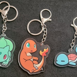 Könnte beinhalten: Drei Schlüsselanhänger mit Comicfiguren aus dem Pokémon-Franchise. Die Schlüsselanhänger sind aus klarem Acryl und zeigen einen grünen Bisasam, einen orangefarbenen Glurak und einen blauen Schiggy.