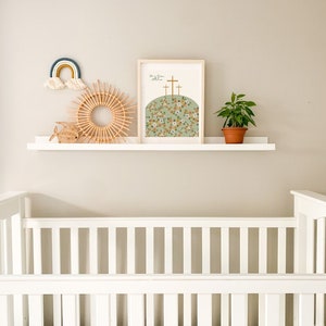 K&ouml;nnte beinhalten: Ein wei&szlig;es Babybett mit einem wei&szlig;en Regal dar&uuml;ber. Das Regal ist mit einem Regenbogen-Wandbehang, einem gewebten sonnenf&ouml;rmigen Wandbehang, einem kleinen Holzfisch und einem gerahmten Druck mit dem Text "Er ist auferstanden" und einer floralen Illustration von drei Kreuzen auf einem H&uuml;gel dekoriert.
