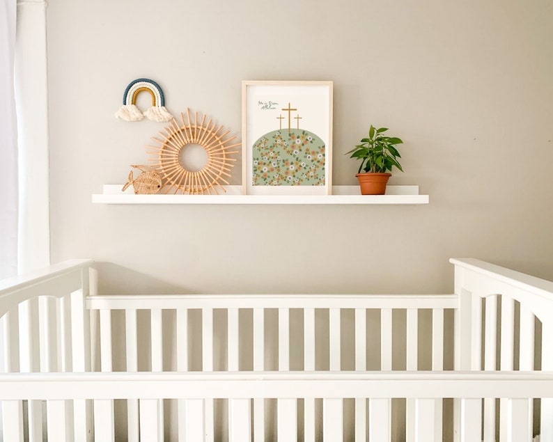 K&ouml;nnte beinhalten: Ein wei&szlig;es Babybett mit einem wei&szlig;en Regal dar&uuml;ber. Das Regal ist mit einem Regenbogen-Wandbehang, einem gewebten sonnenf&ouml;rmigen Wandbehang, einem kleinen Holzfisch und einem gerahmten Druck mit dem Text "Er ist auferstanden" und einer floralen Illustration von drei Kreuzen auf einem H&uuml;gel dekoriert.