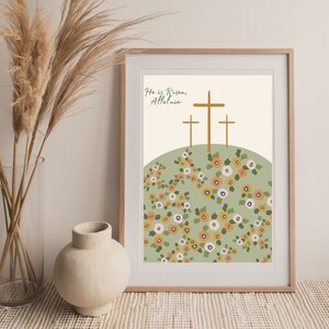 K&ouml;nnte beinhalten: Ein digitaler Kunstdruck mit einem gr&uuml;nen H&uuml;gel und drei goldenen Kreuzen. Der Text "He is Risen, Alleluia" ist oben auf dem Druck in einer Schreibschrift geschrieben. Der H&uuml;gel ist mit einem Muster aus orangefarbenen, wei&szlig;en und blauen Blumen bedeckt.