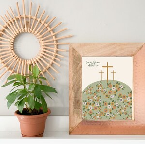 K&ouml;nnte beinhalten: Ein gerahmter Druck mit einem gr&uuml;nen H&uuml;gel und drei goldenen Kreuzen. Der Druck lautet "He is Risen, Alleluia." Der H&uuml;gel ist mit gelben und wei&szlig;en Blumen bedeckt.
