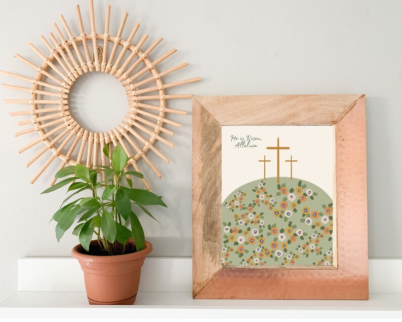 K&ouml;nnte beinhalten: Ein gerahmter Druck mit einem gr&uuml;nen H&uuml;gel und drei goldenen Kreuzen. Der Druck lautet "He is Risen, Alleluia." Der H&uuml;gel ist mit gelben und wei&szlig;en Blumen bedeckt.