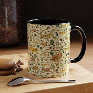 Puede incluir: Una taza de cer&aacute;mica con asa e interior negros. El exterior presenta un fondo beige con un patr&oacute;n repetido de lagartos de dibujos animados, cactus y otras plantas del desierto. Una cuchara y un pastel est&aacute;n en primer plano.