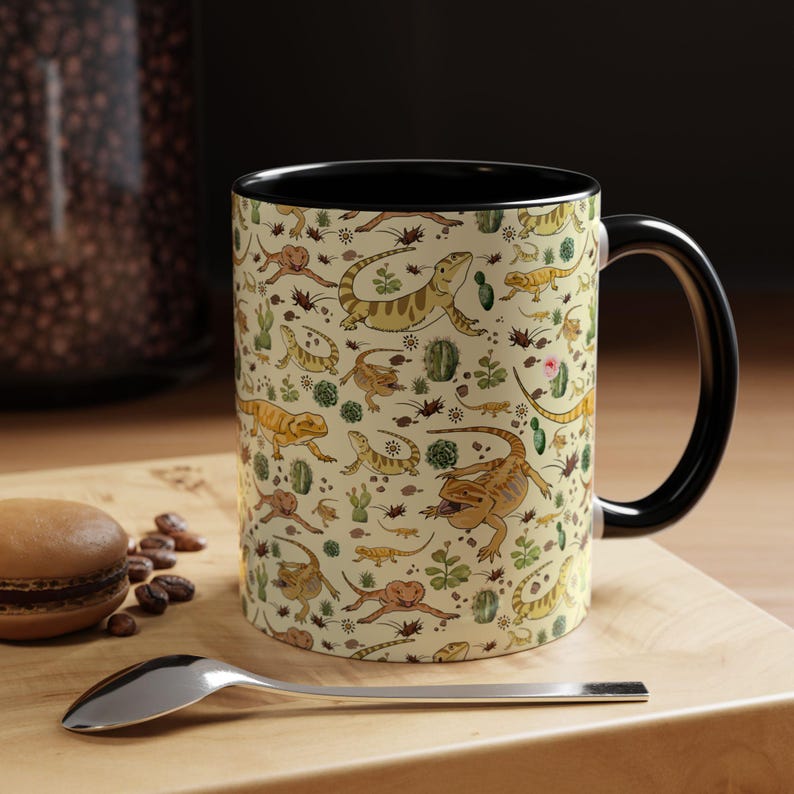 Puede incluir: Una taza de cer&aacute;mica con asa e interior negros. El exterior presenta un fondo beige con un patr&oacute;n repetido de lagartos de dibujos animados, cactus y otras plantas del desierto. Una cuchara y un pastel est&aacute;n en primer plano.