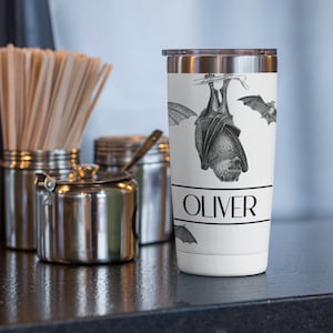 May include: A white stainless steel tumbler with a black and white illustration of a bat hanging upside down. The tumbler has the name "Oliver" printed on the front.