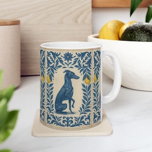 May include: White ceramic mug featuring a blue illustration of a greyhound dog within a decorative blue and cream border. The design includes lemons and floral elements. The mug sits on a beige coaster.