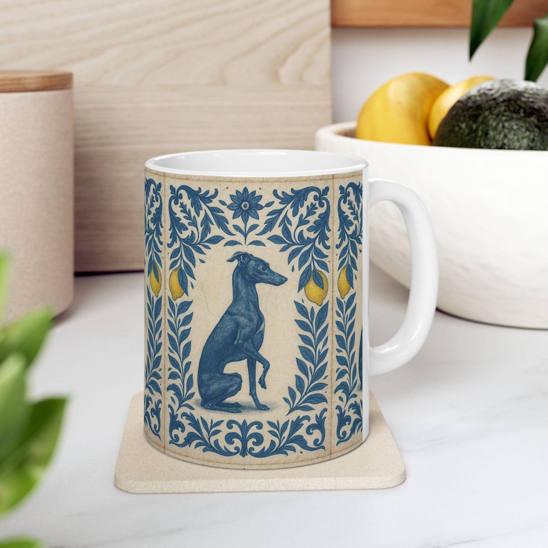 May include: White ceramic mug featuring a blue illustration of a greyhound dog within a decorative blue and cream border. The design includes lemons and floral elements. The mug sits on a beige coaster.