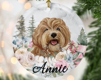 May include: Round ceramic Christmas ornament featuring a smiling brown dog with the name "Annie" in script. The dog is surrounded by watercolor flowers in shades of pink, white, and blue, with a backdrop of trees.