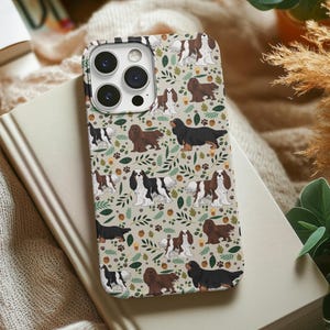 May include: A phone case with a pattern of Cavalier King Charles Spaniels on a beige background with brown leaves and acorns. The case has a camera lens cutout and a black border.