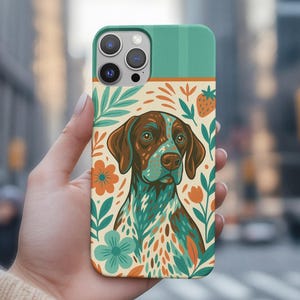 May include: A phone case with a dog illustration. The case has a teal top and a cream background with floral and leaf designs. The dog is brown and teal, with a detailed face. The case is held in a hand.