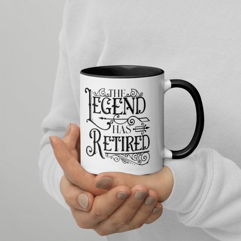 Peut inclure: Une tasse &agrave; caf&eacute; en c&eacute;ramique blanche avec un bord et une poign&eacute;e noirs. La tasse pr&eacute;sente un motif noir qui dit "The Legend Has Retired" avec des fioritures d&eacute;coratives.