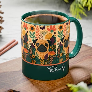 May include: A ceramic mug with a green handle and rim, featuring a pug design amidst colorful flowers and foliage. The mug has a dark green base with the name "Emily" in cursive. The mug is filled with a dark liquid.