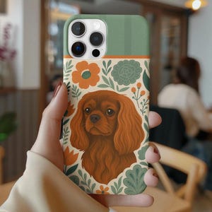 May include: A phone case featuring a Cavalier King Charles Spaniel illustration. The case has a green top, a floral design with orange and green flowers, and a brown dog portrait. The case is held in a hand.