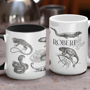 May include: White ceramic mug with a black interior and handle, decorated with grayscale illustrations of reptiles. The mug on the right has the name "ROBERT" printed in black. A unique gift for reptile lovers.