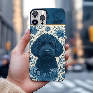 May include: A navy blue phone case featuring a detailed illustration of a black dog with curly fur, surrounded by blue floral patterns on a cream background. The top section has a blue design. The case is held in a hand.