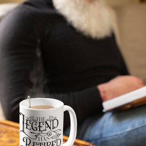 Peut inclure: Une tasse &agrave; caf&eacute; en c&eacute;ramique blanche avec du texte noir qui dit "The Legend Has Retired". La tasse est pos&eacute;e sur une surface en bois. Une personne est assise &agrave; l'arri&egrave;re-plan, floue.