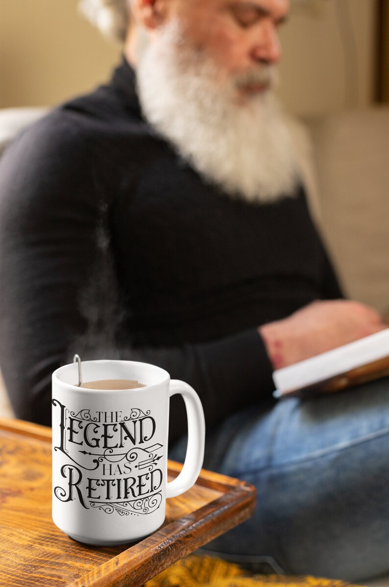 Peut inclure: Une tasse &agrave; caf&eacute; en c&eacute;ramique blanche avec du texte noir qui dit "The Legend Has Retired". La tasse est pos&eacute;e sur une surface en bois. Une personne est assise &agrave; l'arri&egrave;re-plan, floue.