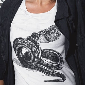 May include: White t-shirt featuring a detailed black and white illustration of a snake coiled around a bird. The snake's scales and the bird's feathers are intricately depicted. The shirt is worn with a black jacket.