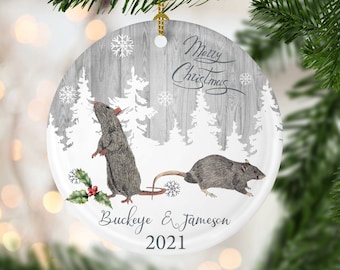 May include: A round Christmas ornament with a gray wood grain background and a winter forest scene. Two gray rats are featured, along with the text "Merry Christmas", the names "Buckeye & Jameson", and the year "2021".
