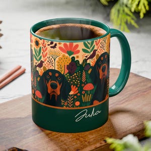 May include: A ceramic coffee mug with a dark green handle and rim. The mug features a colorful floral design with two black and tan dogs. The name "Julia" is written in cursive on the mug.
