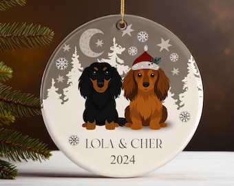 May include: A round ceramic ornament featuring two dachshund dogs. One is black, the other is brown and wearing a Santa hat. The ornament has a winter scene with snowflakes, trees, and the text "LOLA & CHER 2024".