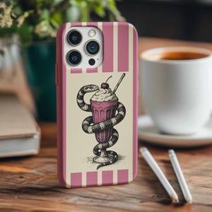 Peut inclure: Coque de t&eacute;l&eacute;phone rose avec un fond cr&egrave;me. Elle pr&eacute;sente un dessin d'un milkshake entour&eacute; d'un serpent, surmont&eacute; d'une cerise. L'&eacute;tui a des rayures verticales roses.