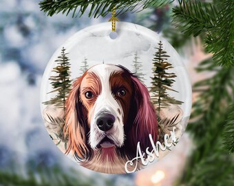 May include: Round ceramic Christmas ornament featuring a dog's portrait with the name "Asher" in white script. The dog has brown and white fur, with a forest scene in the background. A gold-colored string is attached for hanging.