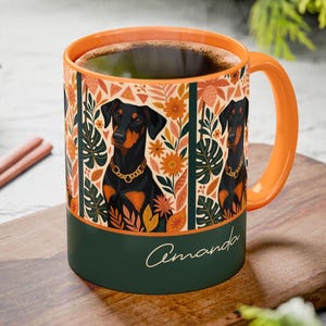 May include: Orange and green ceramic mug featuring a Doberman Pinscher illustration with a gold chain collar. The mug has a floral pattern with the name "Amanda" in a script font on the base. The handle and rim are orange.