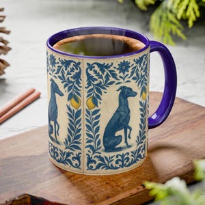 May include: A ceramic mug with a blue handle and rim, featuring a design of blue greyhounds, lemons, and floral patterns on a cream background. The mug contains a dark liquid, possibly coffee or tea.