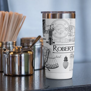 May include: A white stainless steel tumbler with a black and white design featuring a vintage engineering diagram and the name "ROBERT" printed on the side.