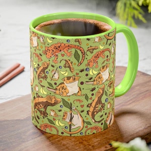 May include: A ceramic mug with a green interior and handle, filled with a dark liquid. The exterior features a repeating pattern of orange and brown geckos, leaves, and fruit on a green background. The mug is sitting on a wooden surface.