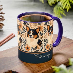Puede incluir: Una taza de cerámica con asa y borde morados, con un diseño de corgi entre patrones florales. La taza tiene una base azul marino con el nombre "Emmy" en cursiva. La taza está llena de un líquido oscuro.