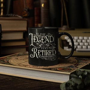 Peut inclure: Une tasse &agrave; caf&eacute; noire avec du texte blanc qui dit "The Legend Has Retired". La tasse est pos&eacute;e sur un livre avec un motif de carte.