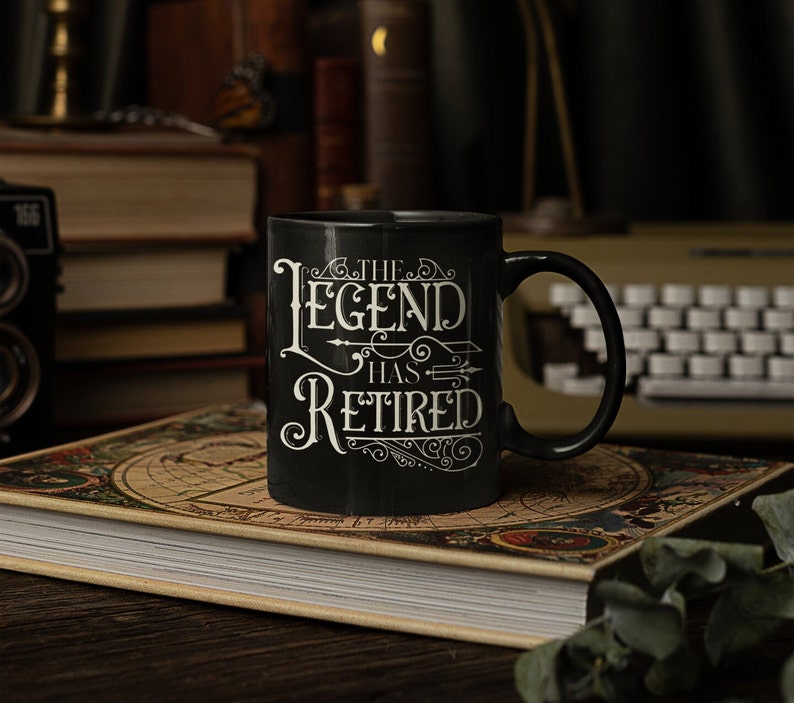 Peut inclure: Une tasse &agrave; caf&eacute; noire avec du texte blanc qui dit "The Legend Has Retired". La tasse est pos&eacute;e sur un livre avec un motif de carte.