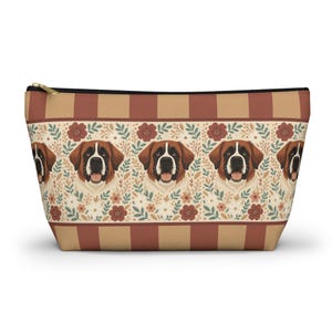 May include: A zippered cosmetic bag featuring a repeating pattern of Saint Bernard dog faces with brown and white fur, set against a floral background. The top and bottom borders have a striped design in shades of brown and tan.