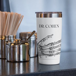 May include: White stainless steel travel mug with a black and white anatomical hand illustration and the text "DR. COHEN" printed on the side.