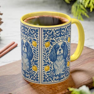 May include: A ceramic mug with a yellow handle and rim, filled with a dark liquid. The mug's exterior features a blue and white floral design with a Cavalier King Charles Spaniel and lemons. The mug sits on a wooden surface.
