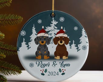 May include: Round Christmas ornament featuring two dachshund dogs wearing Santa hats. The ornament has a teal background with white snowflakes and trees. The names "Roger & Luna" and the year "2024" are printed on the ornament.