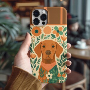 May include: A phone case with a brown dog illustration and floral design. The case has a brown top and a floral pattern with orange, green, and white flowers. The dog wears a red-orange bandana. The case is held in a person's hand.
