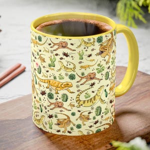 Puede incluir: Una taza de caf&eacute; amarilla con asa y borde amarillos, llena de l&iacute;quido oscuro. La taza presenta un patr&oacute;n repetido de lagartos de dibujos animados, cactus e insectos sobre un fondo crema. La taza est&aacute; sobre una superficie de madera.