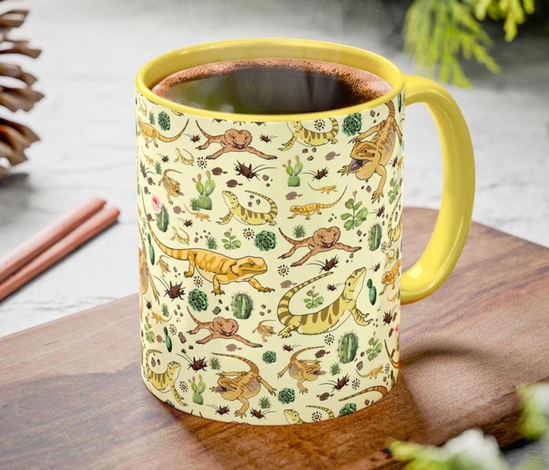 Puede incluir: Una taza de caf&eacute; amarilla con asa y borde amarillos, llena de l&iacute;quido oscuro. La taza presenta un patr&oacute;n repetido de lagartos de dibujos animados, cactus e insectos sobre un fondo crema. La taza est&aacute; sobre una superficie de madera.