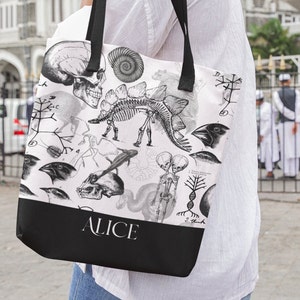 May include: A white tote bag with black straps and a black bottom. The bag has a white background with black and white illustrations of anatomical and scientific imagery, including a dinosaur skeleton, a human skull, and a bird. The text "ALICE" is printed in black on the bottom of the bag.