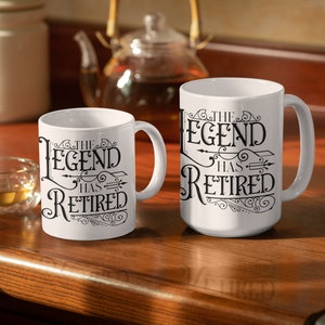 Peut inclure: Deux tasses en c&eacute;ramique blanche avec du texte noir qui dit "The Legend Has Retired" sur une surface en bois. Les tasses sont de tailles diff&eacute;rentes.