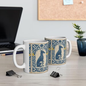 May include: Two white ceramic mugs with a blue and yellow design. The design features a blue dog, surrounded by blue leaves and yellow lemons. The mugs are on a light wood surface.