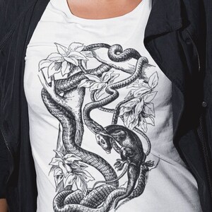 May include: White t-shirt featuring a detailed black and white illustration of a snake coiled around a tree branch with leaves and a small animal. The design is centered on the shirt.