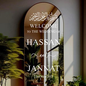 以下が含まれることがあります： アーチ型のトップを持つ金色の鏡で、結婚式のお知らせが書かれています。お知らせには、「Bismillah al-Rahman al-Rahim」、「HassanとJannatの結婚式へようこそ」、「2025年5月23日」と書いてあります。