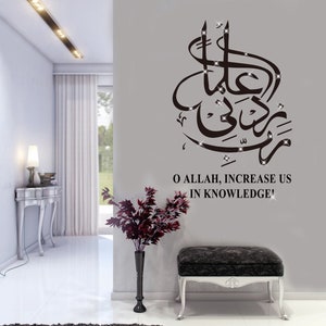 May include: A black and white wall decal with Arabic calligraphy and the text "O Allah, Increase Us In Knowledge!" The decal is on a grey wall in a room with a white table and a vase of red flowers.