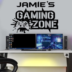 Könnte beinhalten: Eine weiße Wand mit einem schwarzen Vinyl-Aufkleber, auf dem "Jamie's Gaming Zone" mit einer stilisierten Videospiel-Controller-Grafik steht. Zwei schwarze Computermonitore stehen auf einem weißen Schreibtisch mit schwarzen Lautsprechern und einem schwarzen Stuhl.